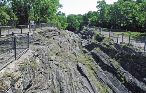 A rocky gorge with steep sides, surrounded by greenery and a fence, with people walking along the paths above.