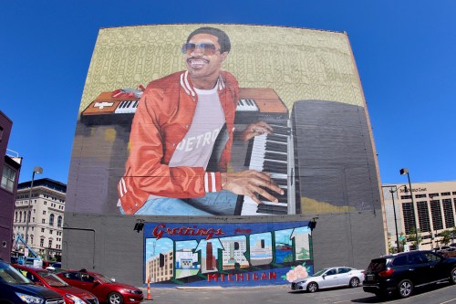 A vibrant mural of a smiling musician playing piano, with "Greetings from Detroit, Michigan" at the bottom.
