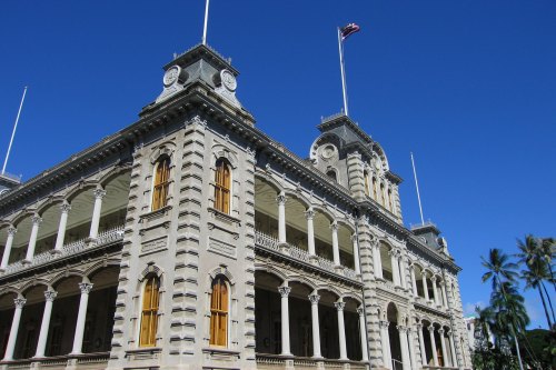Historic building with ornate architecture, tall columns, and a flag flying against a clear blue sky. Palm trees in view.