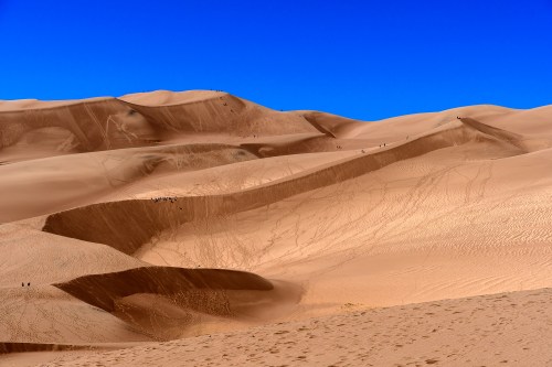 Vast sandy dunes under a clear blue sky, with small figures visible in the distance.