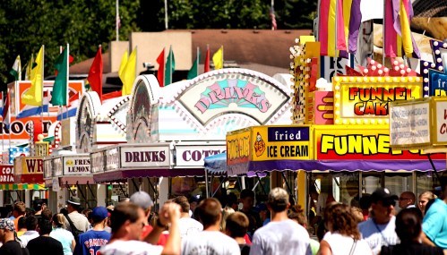 A bustling fair scene with colorful food stalls, flags, and a crowd enjoying various treats like funnel cakes and ice cream.