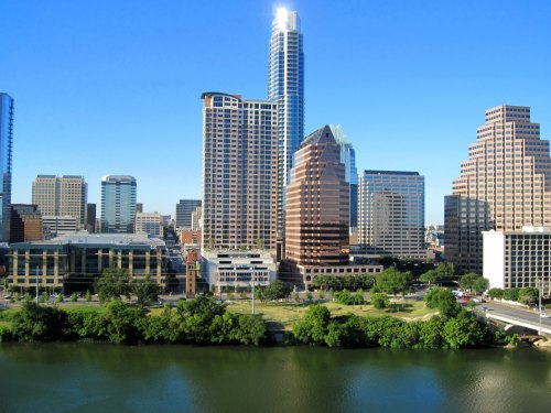 A skyline view of tall buildings along a river, with clear blue skies and green trees in the foreground.