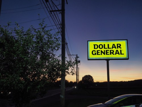 A Dollar General sign illuminated at dusk, with trees and power lines in the foreground against a colorful sunset.