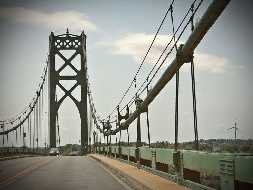 A green suspension bridge spans a road, with wind turbines visible in the background under a cloudy sky.