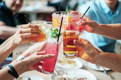 A group of people toasting with colorful cocktails and drinks at a table, surrounded by food.