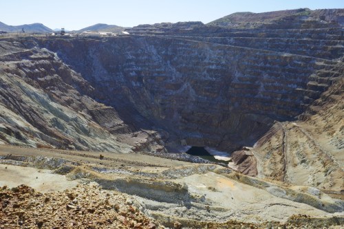 A vast open-pit mine with layered rock formations and a small body of water at the bottom, under a clear blue sky.