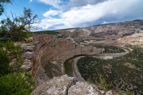 A winding river flows through a canyon surrounded by rocky cliffs and green vegetation under a cloudy sky.