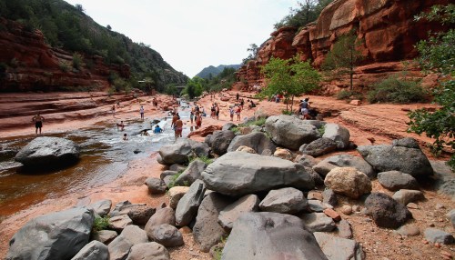 People enjoying a river surrounded by red rock formations and greenery on a sunny day.