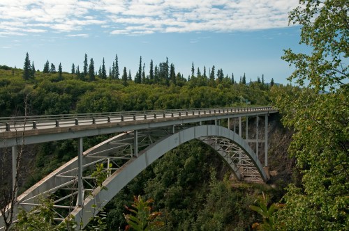 A large, arched bridge spans a lush green valley under a blue sky with scattered clouds.
