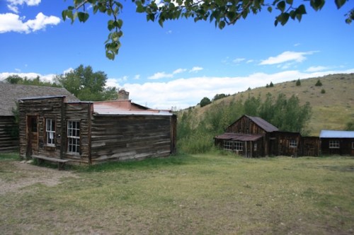 Two rustic wooden buildings sit on grassy land, surrounded by trees and rolling hills under a blue sky with clouds.