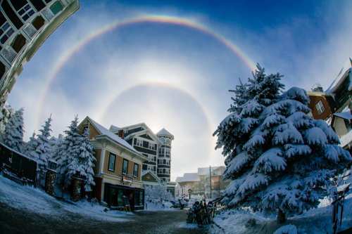 A snowy landscape with frosted trees and buildings, featuring a stunning double rainbow halo in the sky.