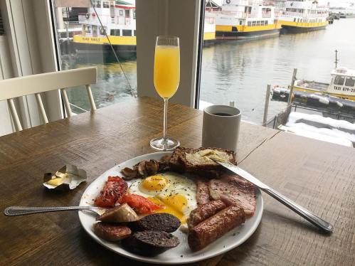 A hearty breakfast plate with eggs, sausages, and toast, alongside a mimosa and coffee, with a waterfront view.
