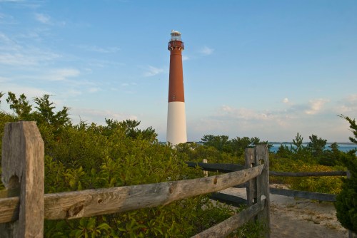 A tall lighthouse stands against a blue sky, surrounded by greenery and a wooden fence in the foreground.