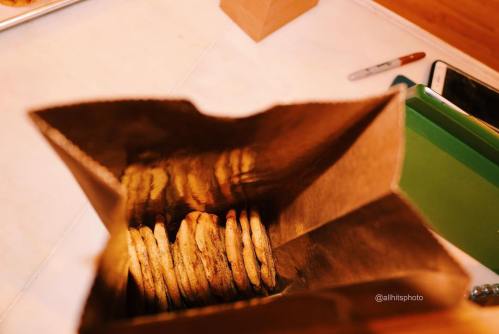 A paper bag filled with freshly baked cookies, viewed from above, with a few items visible in the background.