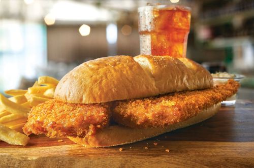 A crispy fish sandwich on a bun, served with fries and a glass of iced tea in a casual dining setting.