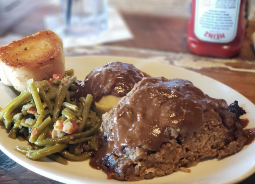 A plate of meatloaf with brown gravy, mashed potatoes, green beans, and a slice of toasted bread.