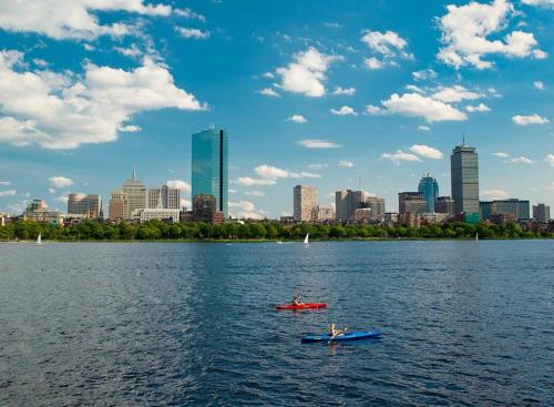 Two kayakers paddle on a calm river with a city skyline and fluffy clouds in the background.