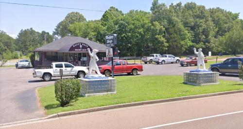 A roadside view of a restaurant with two white statues and parked vehicles in a green area under a clear sky.