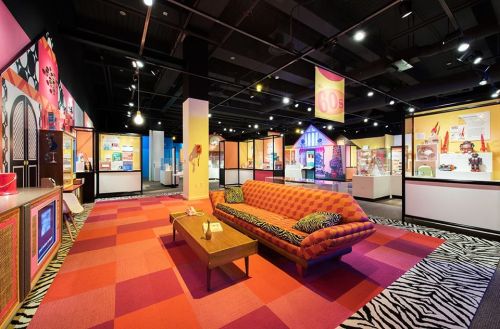 A vibrant museum exhibit featuring a retro 60s living room with a striped rug and colorful displays.