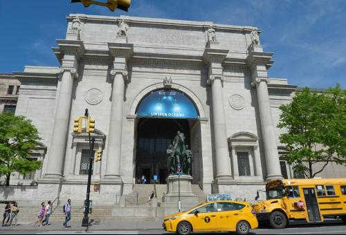 The entrance of a grand museum with a large banner reading "Unseen Oceans," featuring a statue and yellow taxis nearby.
