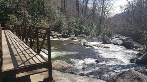 A wooden bridge spans a flowing river, surrounded by trees and rocky banks under a clear blue sky.