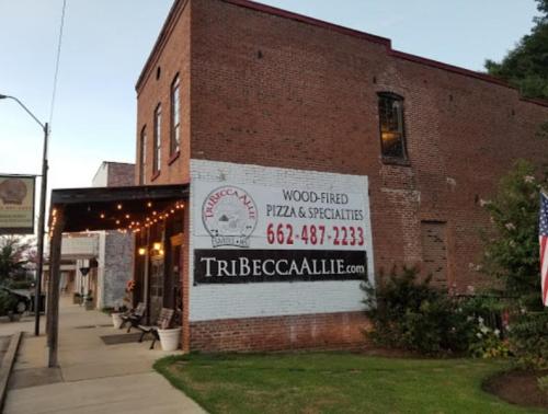 A brick building with a sign for "Tribeca Allie" offering wood-fired pizza and specialties, with outdoor lights.