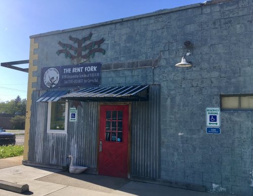 Exterior of The Bent Fork restaurant, featuring a red door, metal siding, and a sign with a logo. Accessible parking signs visible.