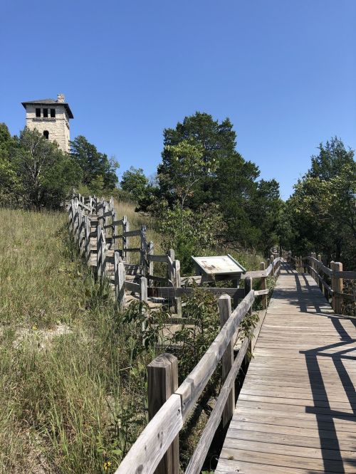 A wooden walkway leads to a stone tower, surrounded by greenery under a clear blue sky.