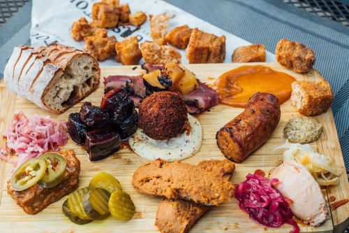 A wooden platter filled with various meats, pickles, bread, and sauces, showcasing a colorful assortment of charcuterie.