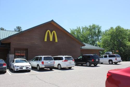 A McDonald's restaurant with a wooden exterior, surrounded by parked cars and trees under a clear blue sky.