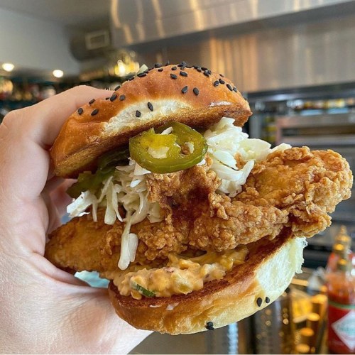 A hand holding a crispy fried chicken sandwich with jalapeños, lettuce, and a spicy sauce on a sesame seed bun.