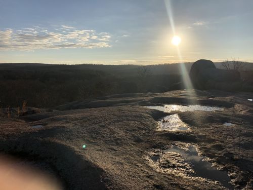 Sunset over a rocky landscape, with puddles reflecting the sky and distant hills under a partly cloudy sky.