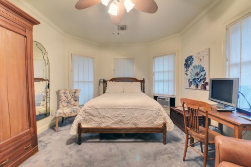 Cozy bedroom featuring a bed, chair, desk, and floral artwork, with natural light from windows.