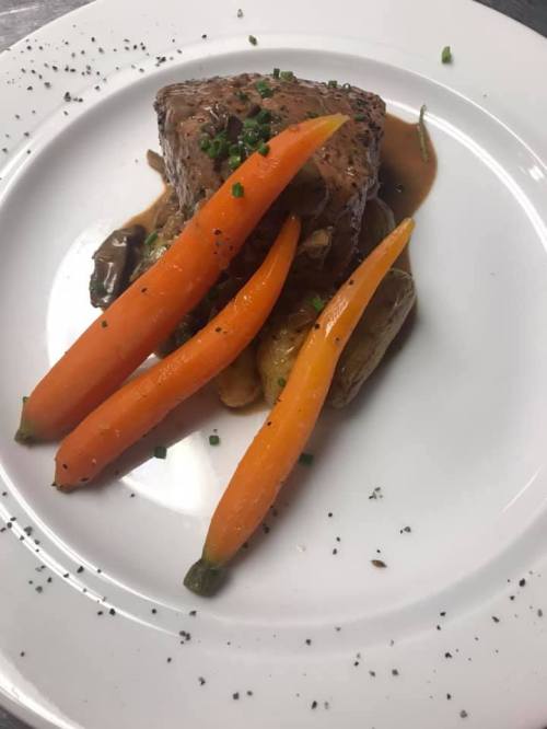 A plated dish featuring a piece of meat, roasted potatoes, and three vibrant orange carrots, garnished with herbs.