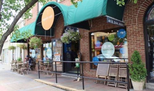 A brick storefront with green awnings, outdoor seating, and colorful decorations in the windows.