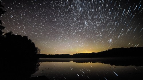 Star trails swirl above a calm lake, reflecting the night sky and surrounding trees in a serene landscape.
