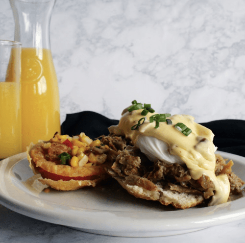 A plate featuring a sandwich with pulled meat, poached egg, and hollandaise sauce, alongside a corn salad and a drink.