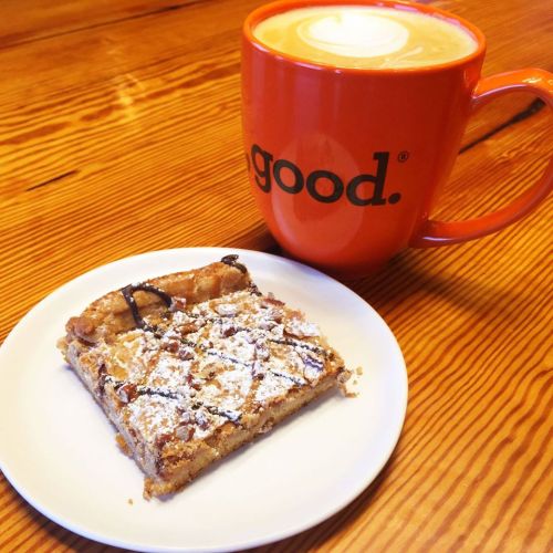 A slice of dessert on a white plate next to a large orange mug of latte on a wooden table.