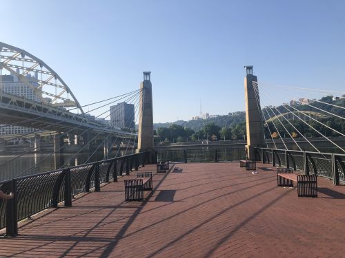 A view of a bridge over a river, with a paved area and benches in the foreground under a clear blue sky.