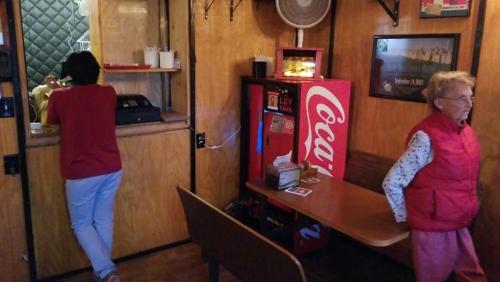 A cozy diner interior with a woman in a red vest, a person at the counter, and a Coca-Cola vending machine.