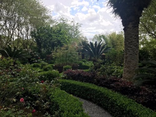 Lush garden with various plants, shrubs, and trees under a partly cloudy sky, featuring winding paths and greenery.