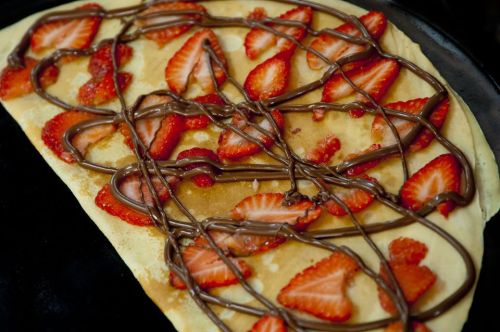 A crepe topped with sliced strawberries and drizzled with chocolate sauce on a black plate.