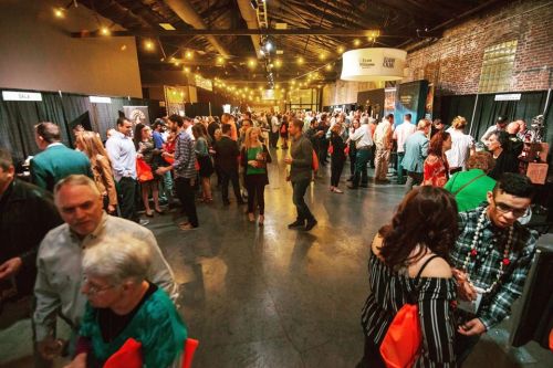 A crowded event space with people mingling, booths set up, and festive lighting overhead.