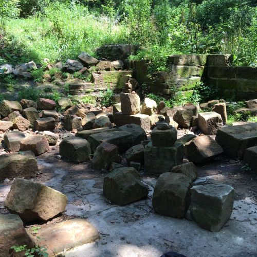 A scattered arrangement of large stones in a green, overgrown area, with trees and foliage in the background.