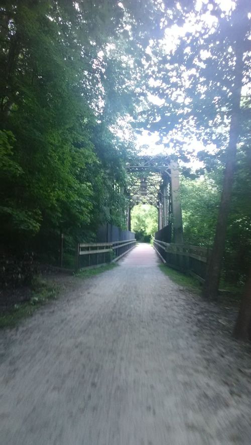A gravel path leads to a green, metal bridge surrounded by lush trees in a serene outdoor setting.