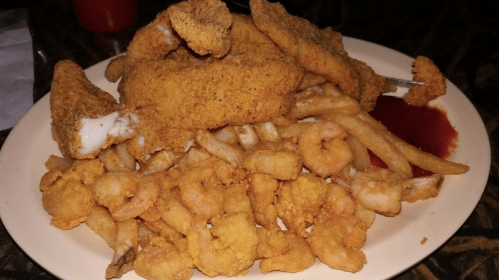 A plate of fried fish fillets, shrimp, and French fries, served with a side of ketchup.