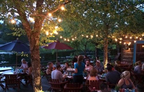 A lively outdoor restaurant scene by a lake, with diners enjoying meals under string lights and shaded umbrellas.