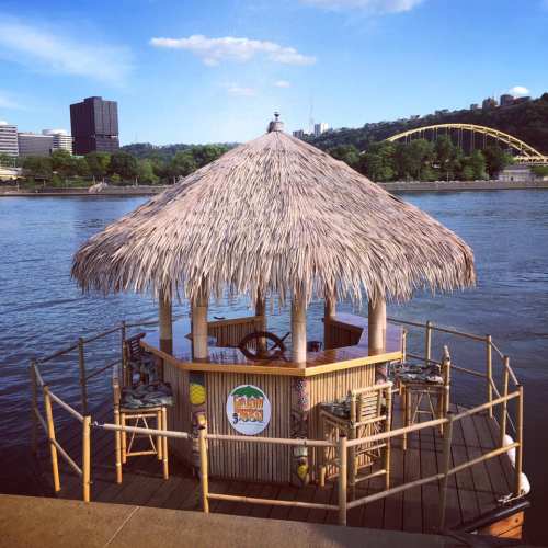 A tiki hut on a river with city buildings in the background and a bridge in the distance under a clear blue sky.