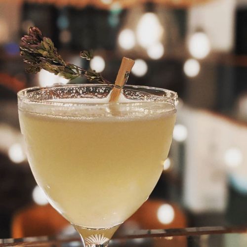 A cocktail in a elegant glass, garnished with a sprig of herbs and a clothespin, set against a blurred background.