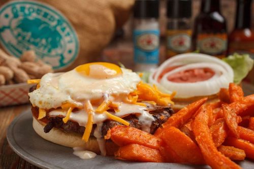 A hearty sandwich topped with a fried egg, cheese, and sauce, served with sweet potato fries and fresh vegetables.